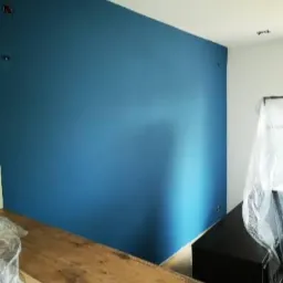 Ściana pomalowana na intensywny odcień niebieskiego, widoczne ślady po mocowaniach, zabezpieczona drewniana półka i meble folią ochronną.
