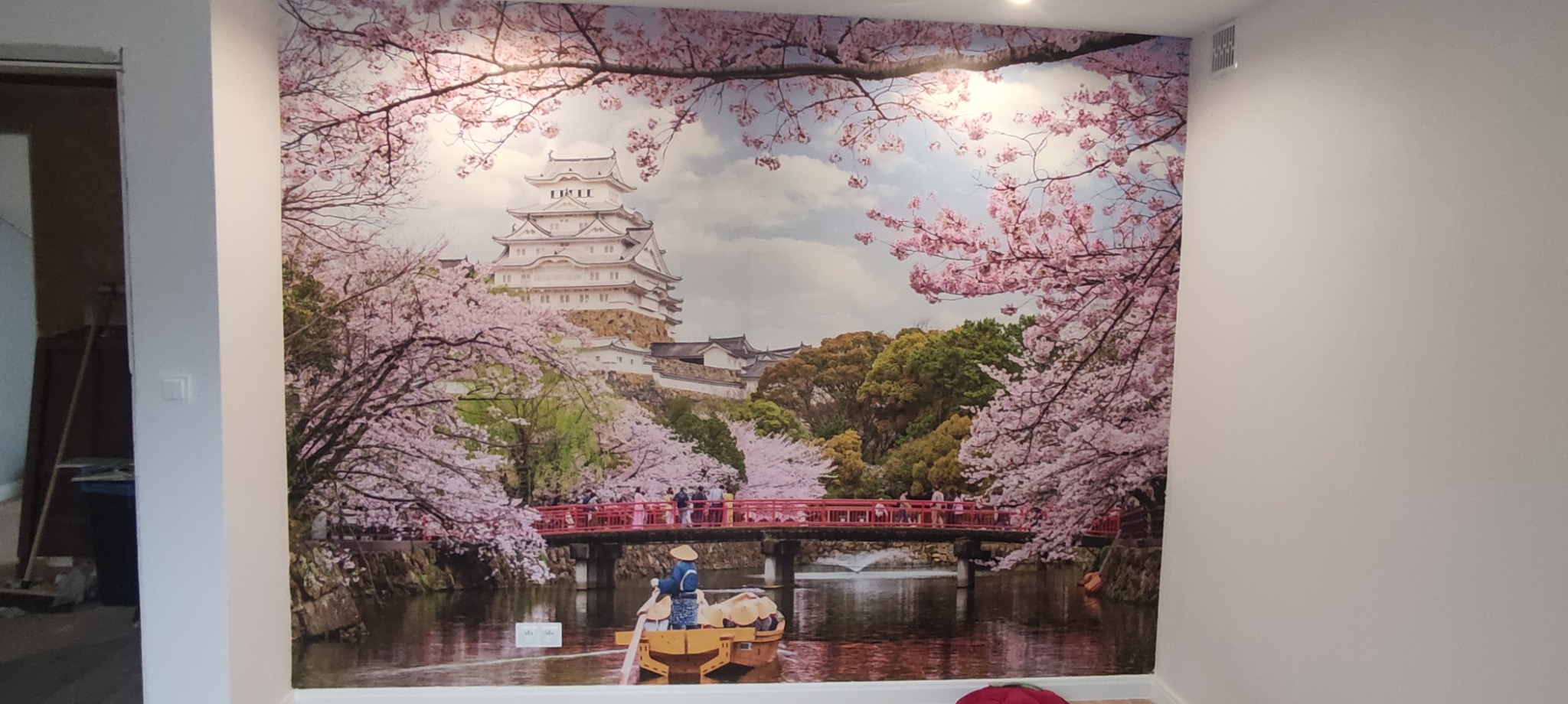 Fototapeta z motywem japońskim: zamek Himeji, kwitnące wiśnie i łódź na rzece, zamontowana na ścianie w jasnym pomieszczeniu.