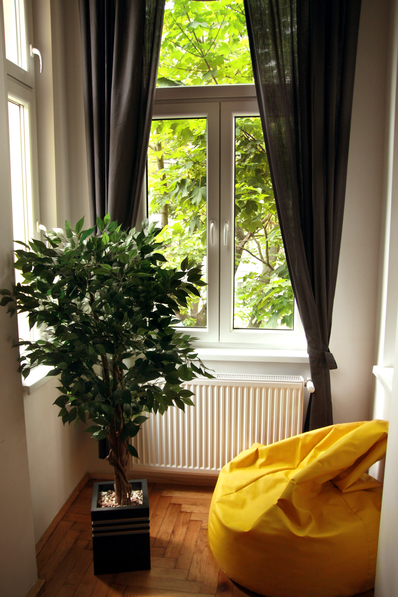 Jasny pokój z oknem wychodzącym na zieleń, zasłoniętym ciemnymi zasłonami, z rośliną doniczkową i żółtą pufą.