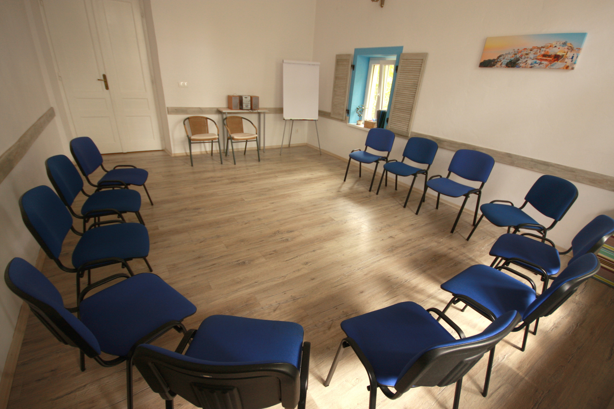 Sala szkoleniowa z krzesłami ustawionymi w okrąg, flipchartem, radiem na stoliku i obrazem Santorini na ścianie.