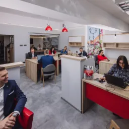 Wnętrze biura agencji nieruchomości w Białymstoku z pracownikami przy biurkach, oświetlone czerwonymi lampami i z logo firmy na ścianie.