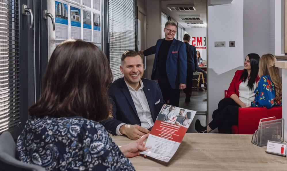 Klientka w biurze nieruchomości otrzymuje ulotkę z ofertą mieszkań od uśmiechniętego agenta, w tle widoczni inni pracownicy i plakaty z wizualizacjami budynków.