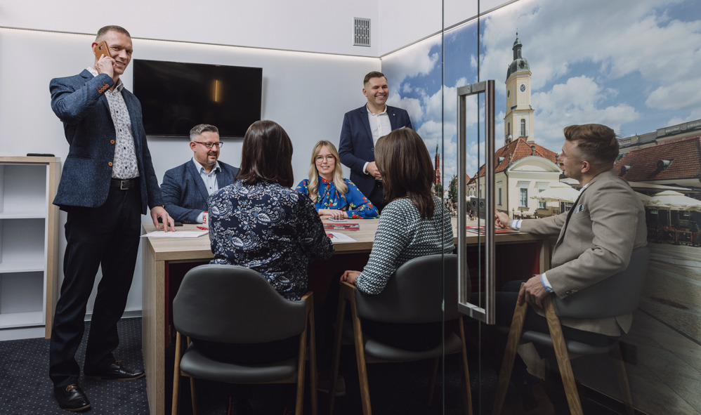 Spotkanie zespołu w biurze z fototapetą przedstawiającą miejski krajobraz, mężczyzna rozmawia przez telefon, pozostali uczestnicy siedzą przy stole.