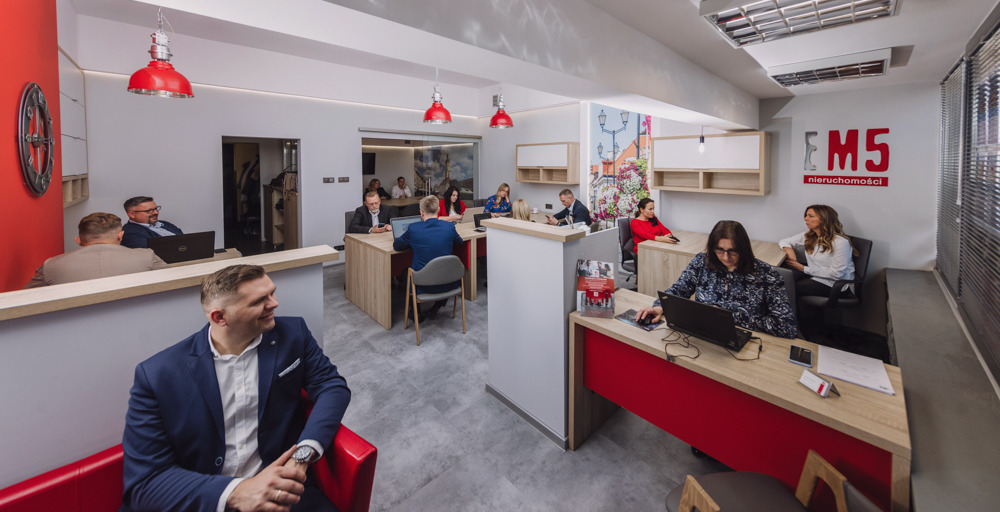Wnętrze biura agencji nieruchomości w Białymstoku z pracownikami przy biurkach, oświetlone czerwonymi lampami i z logo firmy na ścianie.