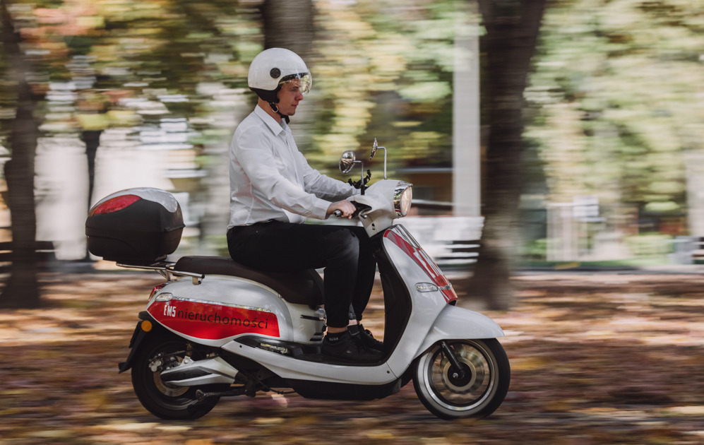 Mężczyzna w kasku na białym skuterze z logo firmy nieruchomości na tle rozmytych drzew.