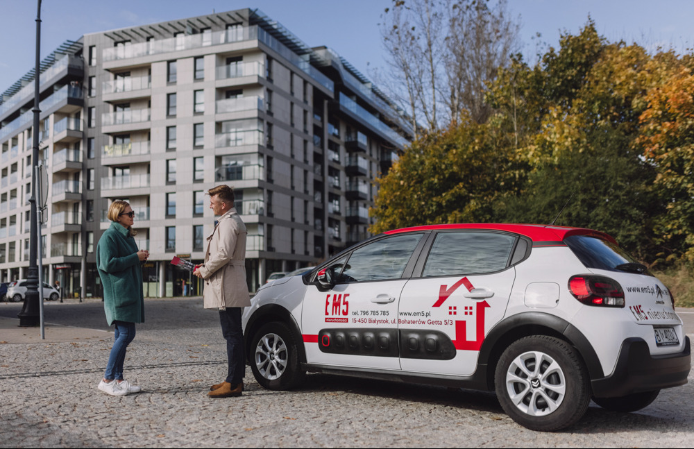 Agent nieruchomości w jasnym płaszczu rozmawia z klientką trzymając w rękach foldery reklamowe na tle nowoczesnego apartamentowca i samochodu firmowego z logo agencji EMS nieruchomości w Białymstoku.