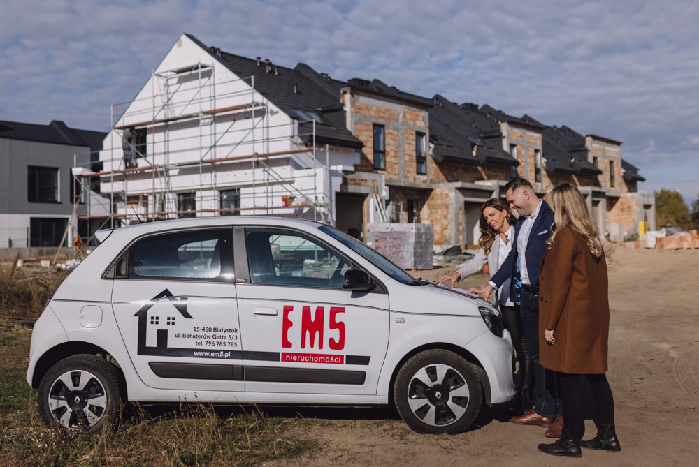 Trzy osoby analizują plany na masce samochodu z logo agencji nieruchomości EM5 na tle budowanych domów szeregowych.