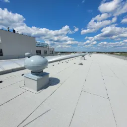 Jasnoszary dach płaski pokryty papą termozgrzewalną z widocznymi kominkami wentylacyjnymi i świetlikami dachowymi, na tle błękitnego nieba z białymi chmurami, w oddali sylwetki dwóch osób...