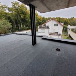 Dream Dach - Czarny, płaski dach pokryty papą termozgrzewalną z widocznymi elementami konstrukcyjnymi i obróbkami blacharskimi na tle zieleni i zabudowań.