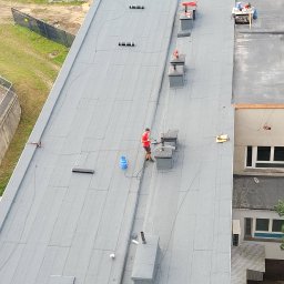 Dream Dach - Praca na płaskim dachu budynku: dekarz w czerwonej koszulce przy kominie, obok butla gazowa i narzędzia. Widok z góry, szary dach, elementy wentylacyjne.