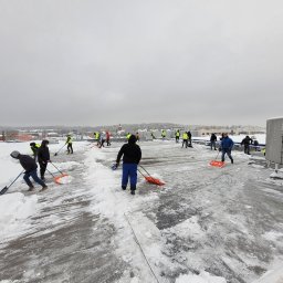 Dream Dach - Grupa pracowników w odblaskowych kamizelkach usuwa śnieg z płaskiego dachu dużego budynku, używając łopat. Część powierzchni jest już oczyszczona, w tle zaśnieżony krajobraz miejski.