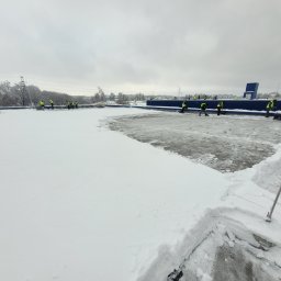 Dream Dach - Zespół pracowników w odblaskowych kurtkach odśnieża duży, płaski dach budynku przemysłowego, usuwając grubą warstwę śniegu łopatami. Widoczna część dachu już oczyszczona.