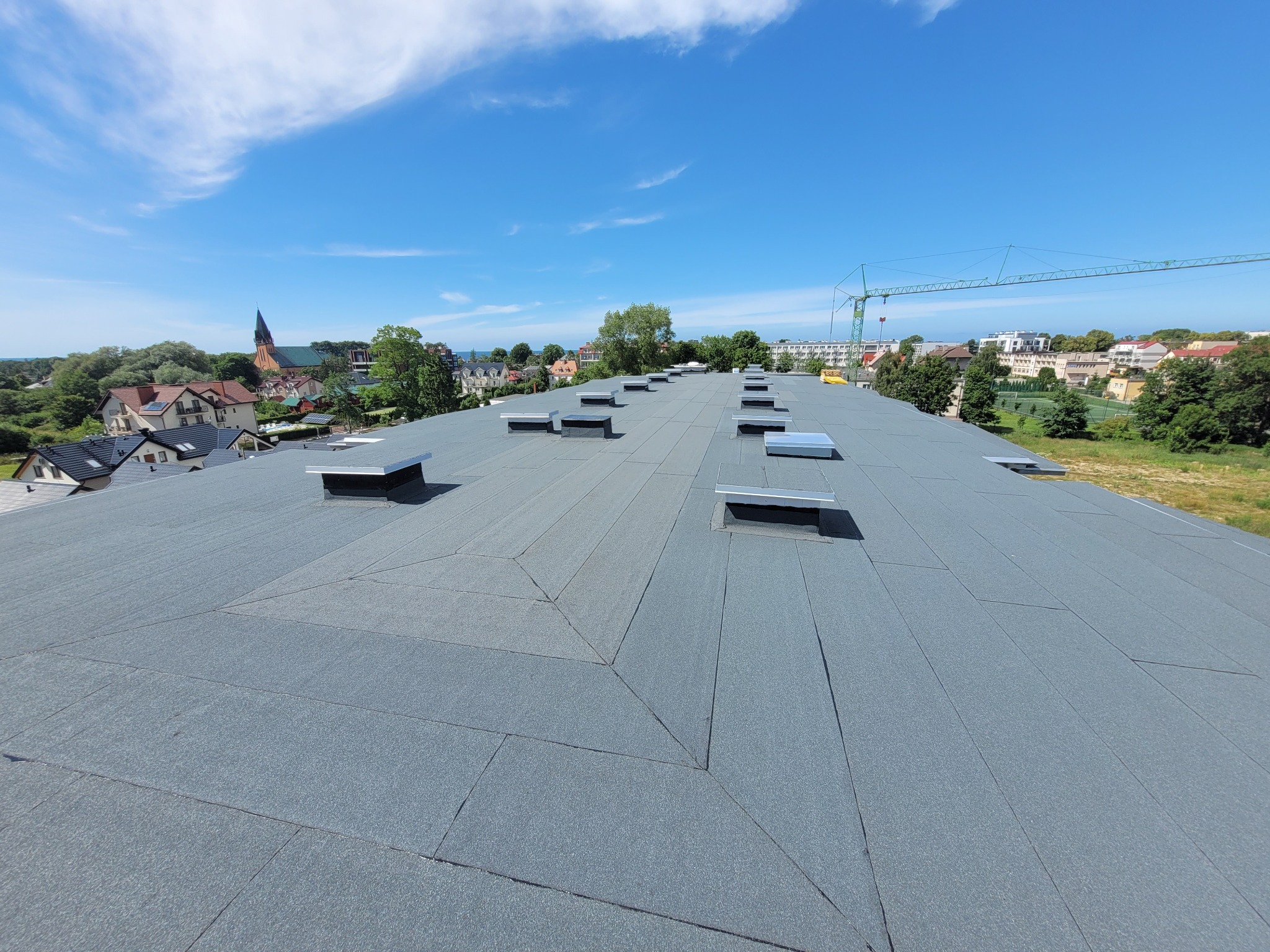 Szary, płaski dach pokryty papą termozgrzewalną z licznymi świetlikami dachowymi, widok na miasto i żuraw budowlany w tle.