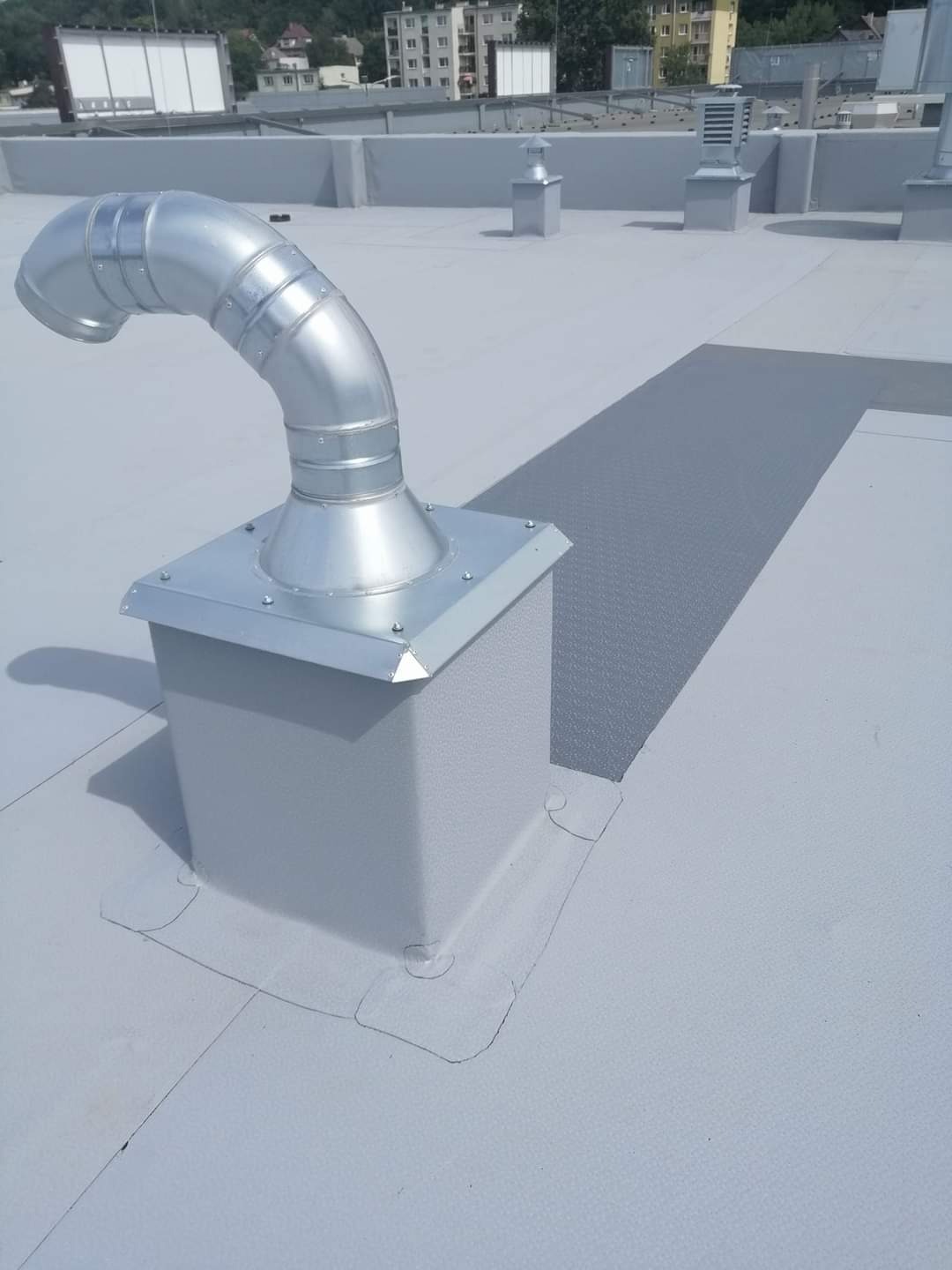 Szary dach płaski z metalową wentylacją kominową i zabezpieczeniem przejścia, widoczne budynki w tle.