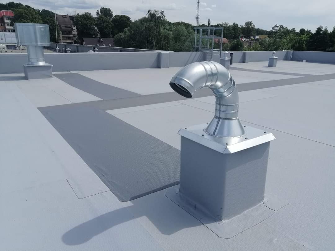 Płaski dach pokryty szarą membraną z widocznymi obróbkami blacharskimi komina wentylacyjnego, w tle zabudowa miejska i zieleń.
