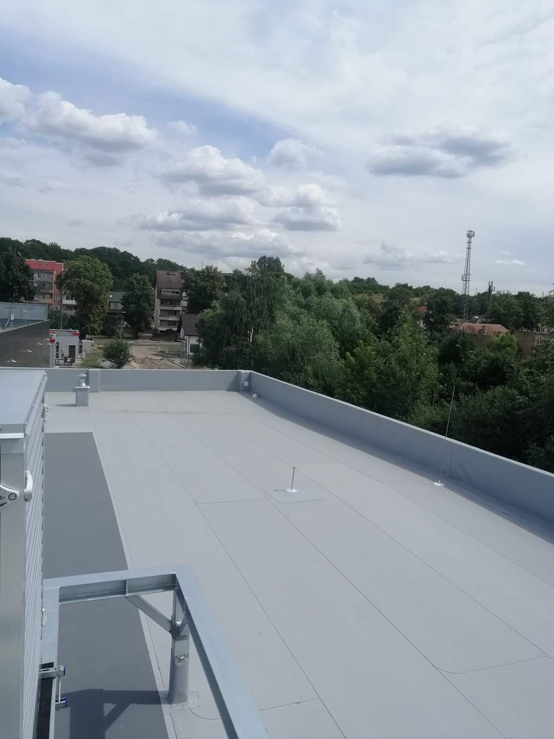 Płaski, szary dach budynku przemysłowego z widocznymi elementami wentylacyjnymi i odgromnikami na tle zieleni i zabudowań mieszkalnych w słoneczny dzień.