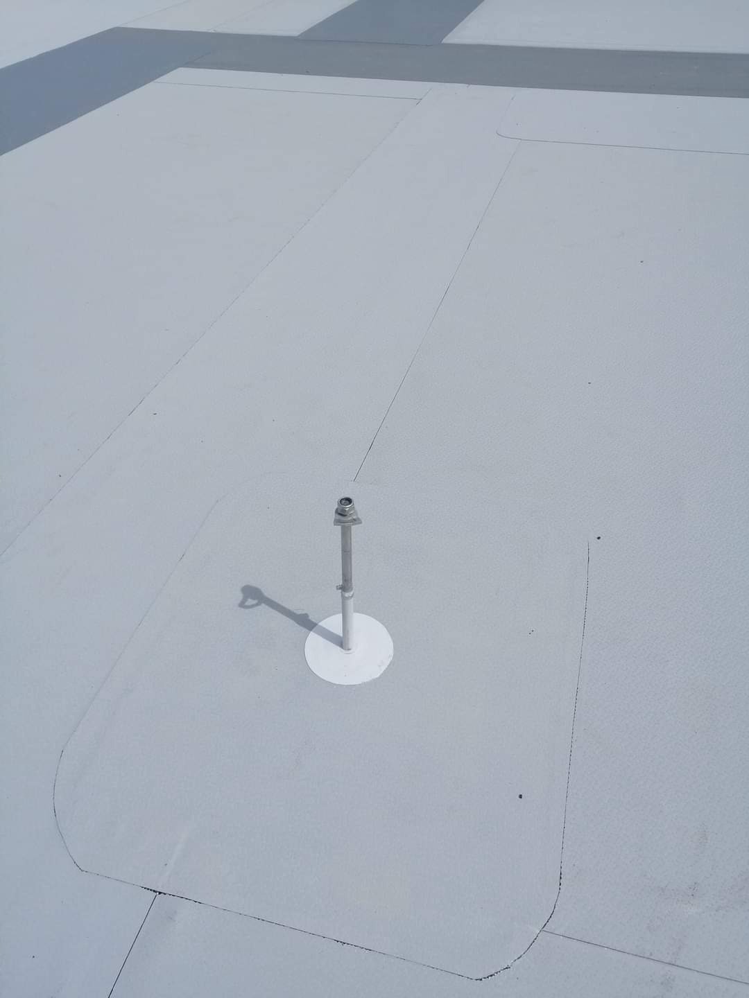 Jasnoszary, płaski dach z widocznym metalowym wspornikiem wystającym z powierzchni, zabezpieczonym białą, okrągłą uszczelką, rzucającym cień w lewo.