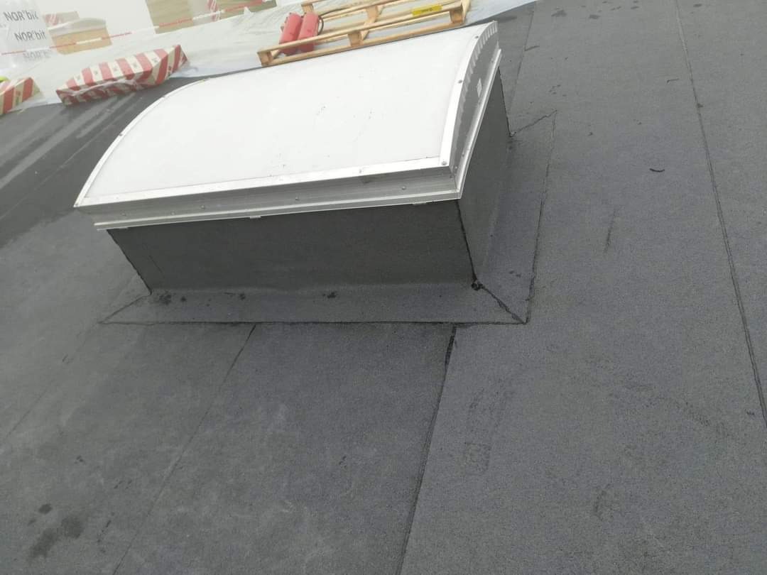 Zamontowany świetlik dachowy na płaskim dachu pokrytym papą, widoczne uszczelnienia i detale obróbki wokół świetlika.