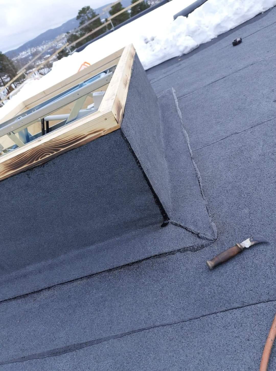 Wykończony fragment dachu z papy z obróbką komina dachowego i narzędziem dekarskim w pobliżu, w tle ośnieżone góry.