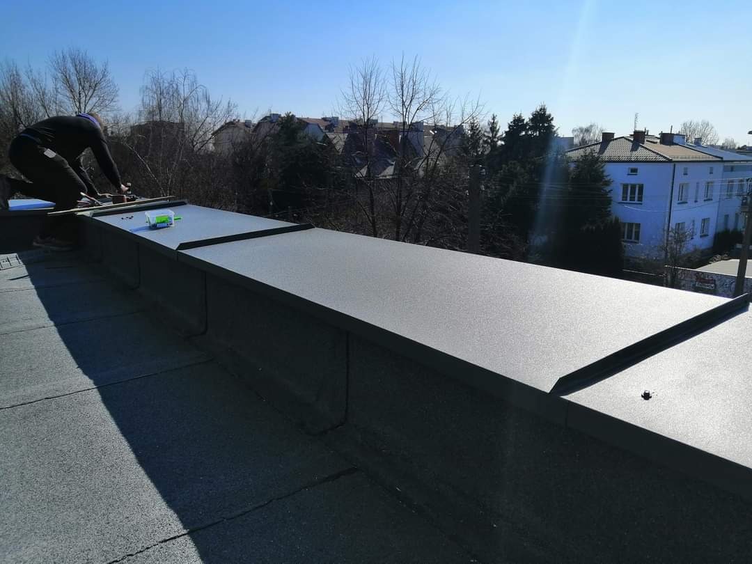 Dekarz montuje obróbki blacharskie na płaskim dachu pokrytym papą, widoczne narzędzia i pudełko z elementami montażowymi, w tle budynki mieszkalne i drzewa bez liści.