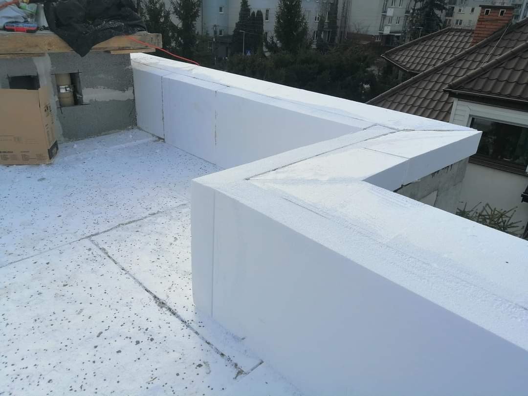 Białe płyty izolacyjne EPS układane na płaskim dachu budynku w trakcie prac budowlanych, widoczne połączenia płyt i fragment muru oraz skrzynka narzędziowa w tle.