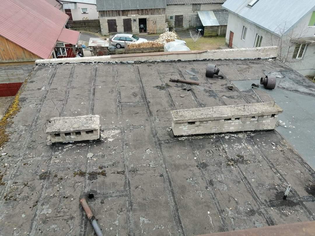 Płaski, zaniedbany dach pokryty papą z betonowymi elementami wentylacyjnymi, widoczne ślady uszkodzeń i zabrudzeń, w tle zabudowania gospodarcze i domy.