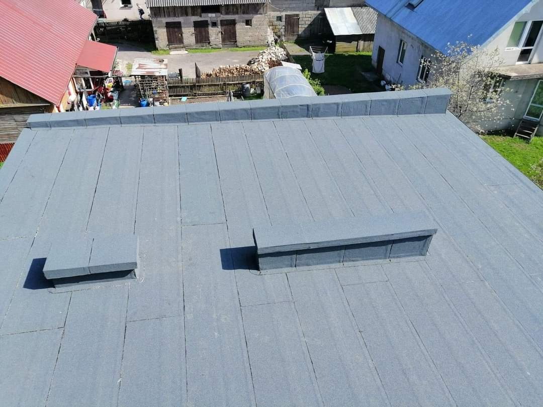 Płaski dach pokryty papą termozgrzewalną z dwoma kominkami wentylacyjnymi, widok z góry na tle zabudowań gospodarczych i domów jednorodzinnych.