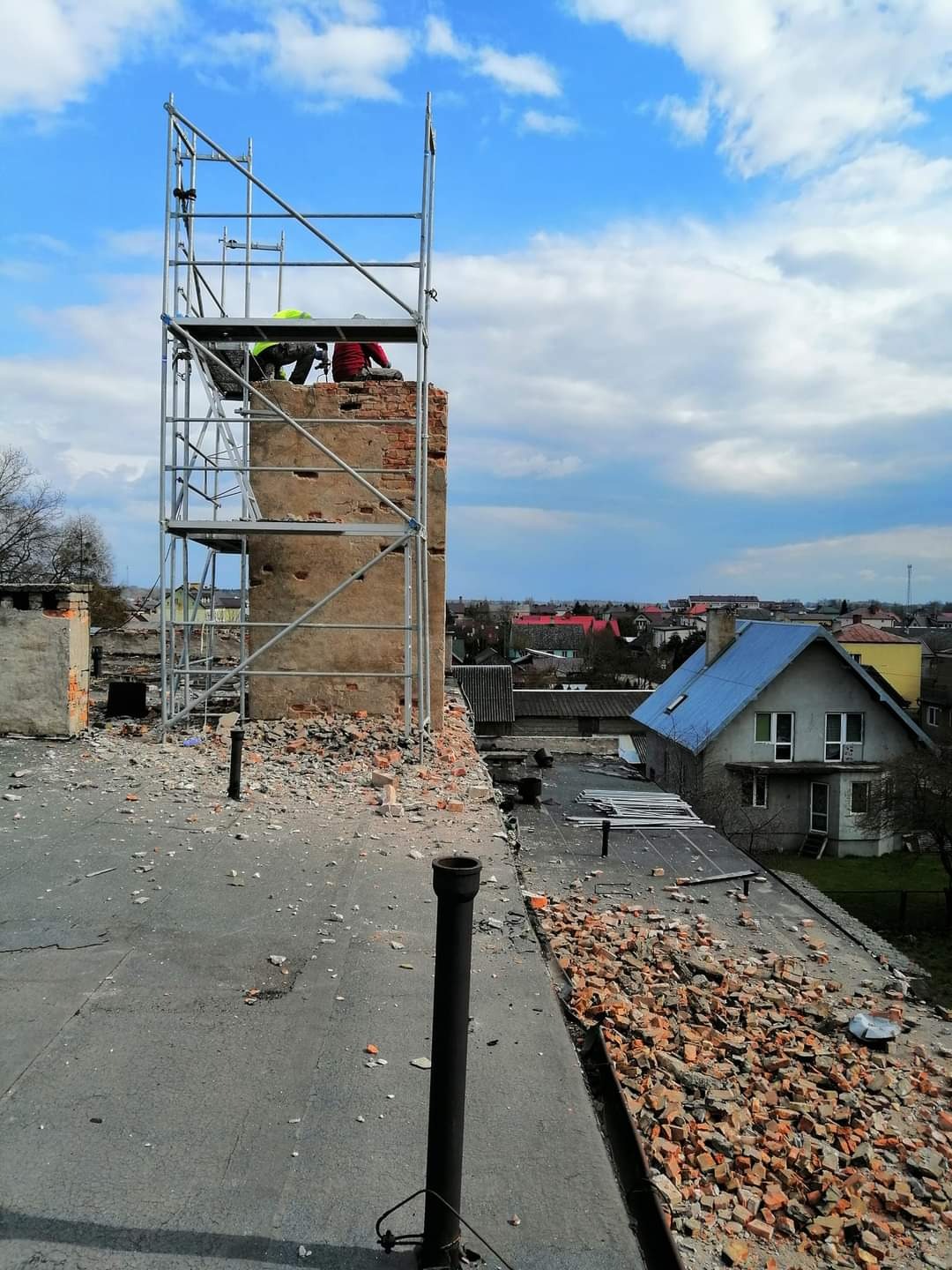 Ekipa dekarzy na rusztowaniu przy ceglanym kominie w trakcie prac na dachu, gruz ceglany na płaskiej powierzchni dachu, widok na zabudowę miejską w tle.
