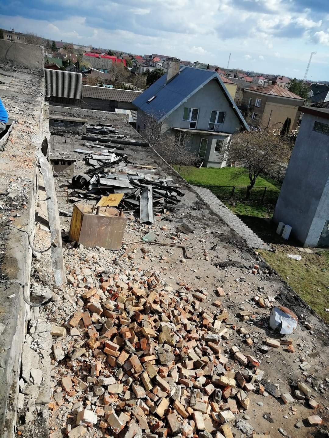 Demontaż dachu: sterta potłuczonych cegieł na płaskim dachu budynku, w tle zabudowa jednorodzinna i pochmurne niebo.