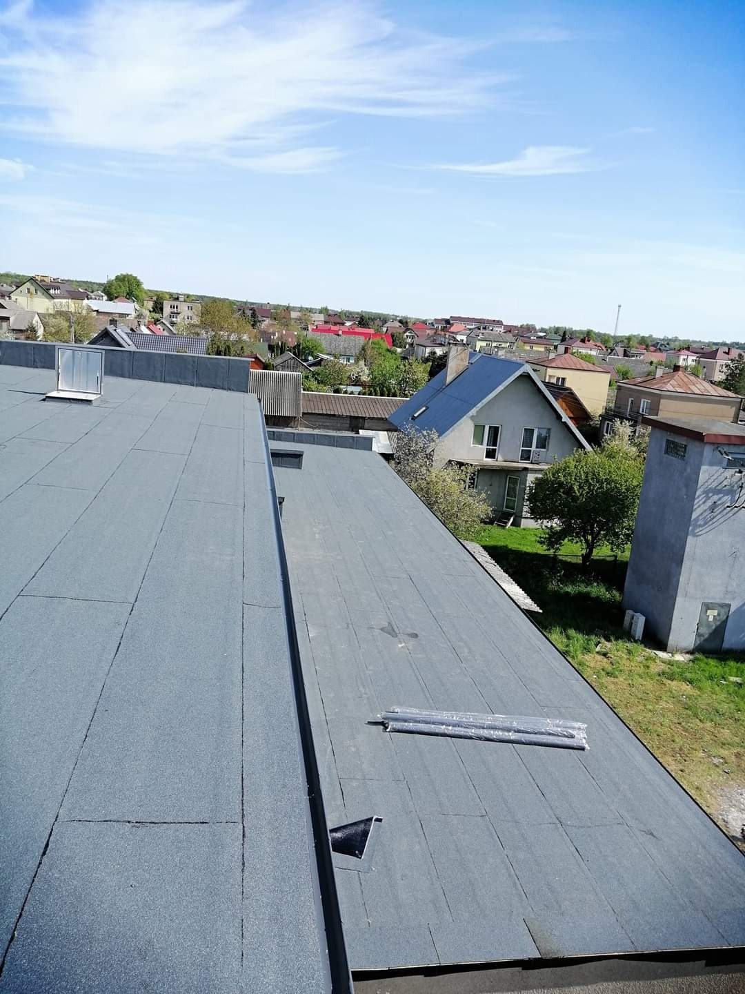 Widok z góry na płaski dach pokryty papą termozgrzewalną z widocznymi zgrzewami, w tle zabudowa jednorodzinna i błękitne niebo.