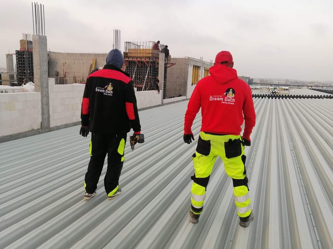 Dwóch dekarzy w firmowych ubraniach z logo 'Dream Dach' stoi na nowym, metalowym dachu z widocznymi szwami, podczas gdy w tle widać konstrukcję budynku w trakcie budowy.