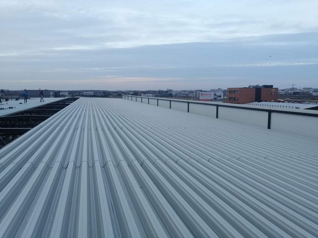 Szeroki, blaszany dach falisty w kolorze srebrnym, z widoczną konstrukcją stalową i ekipą pracowników w oddali, na tle miejskiego krajobrazu pod pochmurnym niebem.