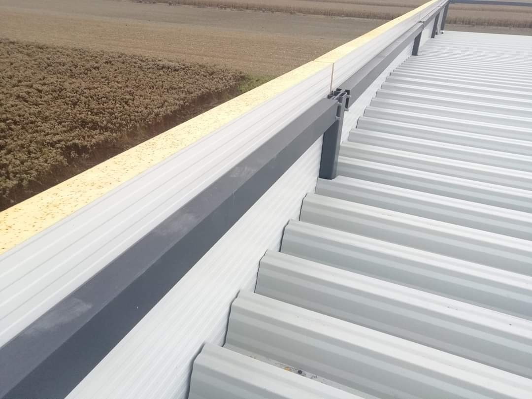 Fragment blaszanej konstrukcji dachowej z szarymi obróbkami blacharskimi i widokiem na pole w tle.