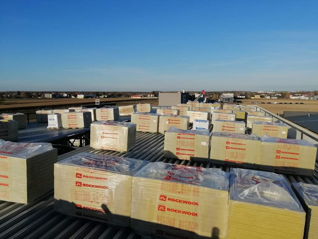 Pakiety wełny mineralnej Rockwool, zabezpieczone folią, ułożone na blaszanym dachu płaskim w słoneczny dzień. W tle zabudowania miejskie i błękitne niebo.