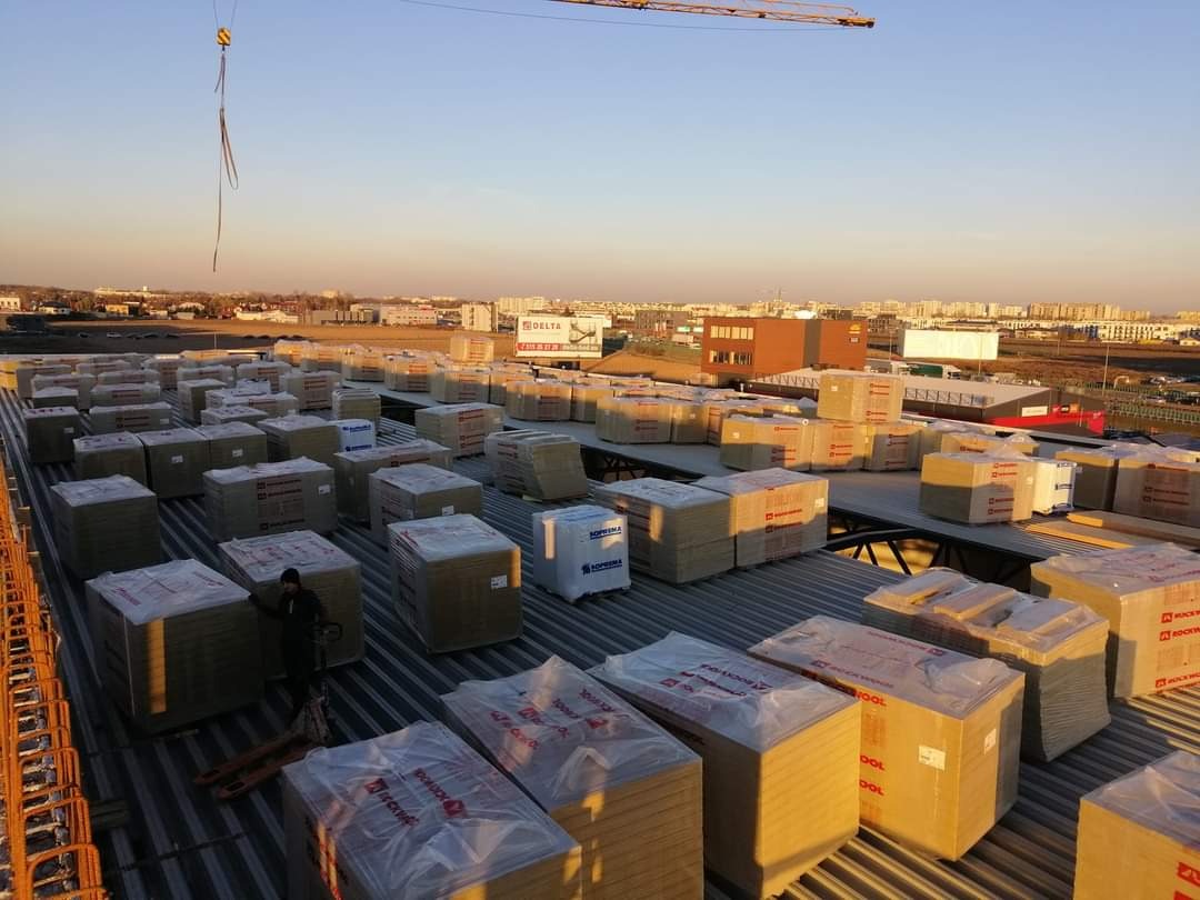 Rozległy dach pokryty licznymi paletami materiałów izolacyjnych, przygotowany do prac budowlanych, w tle widok na miasto i żuraw budowlany.