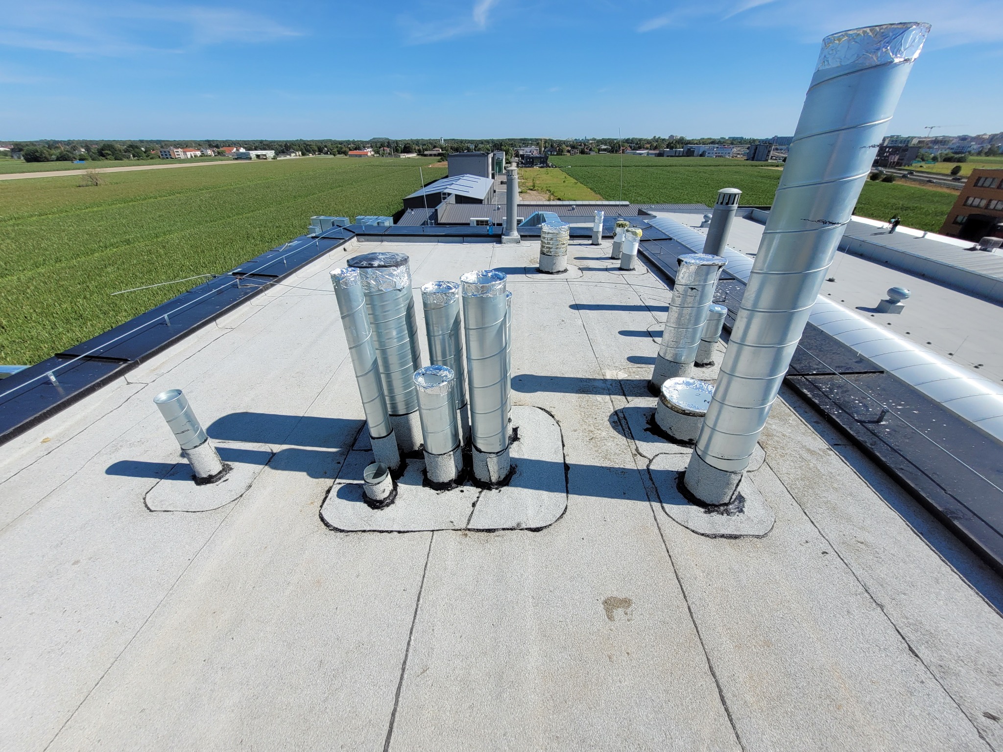 Widok z góry na płaski dach budynku z licznymi, metalowymi kominami wentylacyjnymi, niektóre z izolacją, na tle zielonych pól i błękitnego nieba.