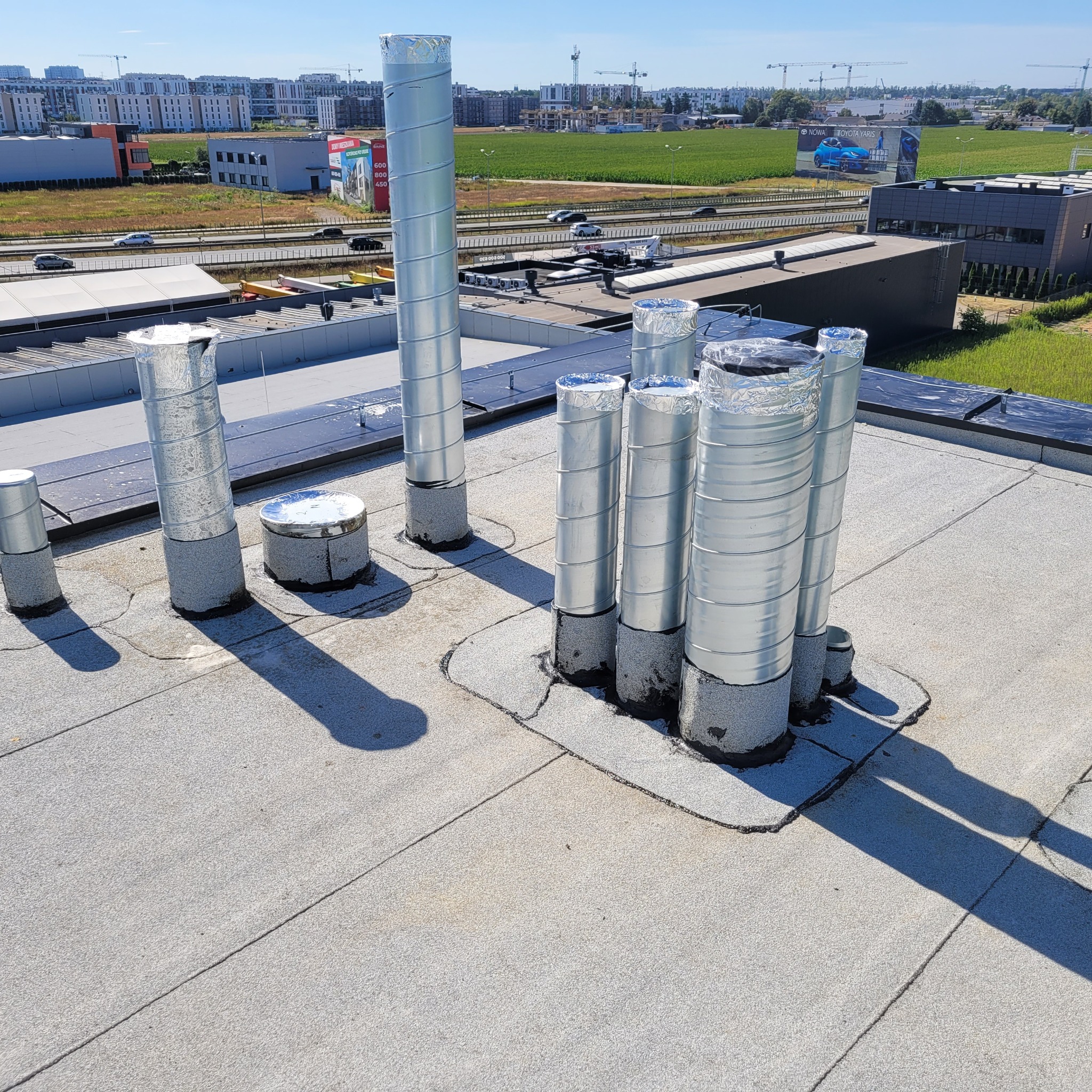 Widok z góry na płaski dach budynku z kilkoma pionowymi, metalowymi rurami wentylacyjnymi o spiralnej strukturze, zabezpieczonymi folią aluminiową na końcach, na tle krajobrazu miejskiego...
