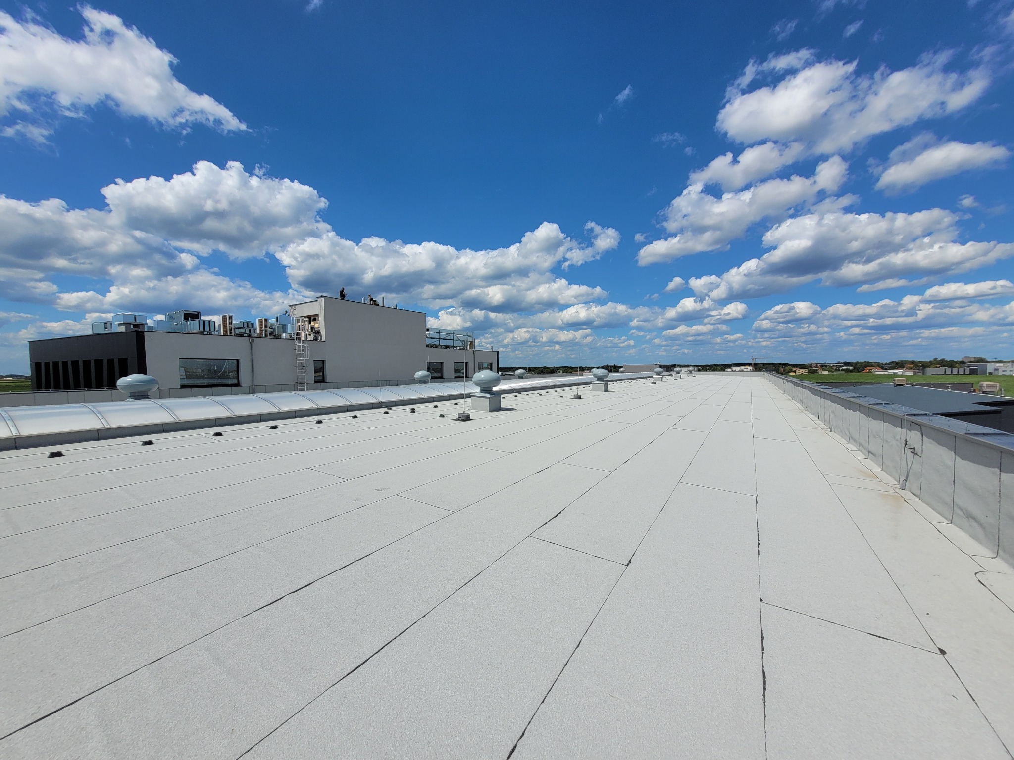 Szeroki, płaski dach przemysłowy pokryty jasną membraną, z widocznymi kominami wentylacyjnymi, budynkami w tle i błękitnym niebem z chmurami.
