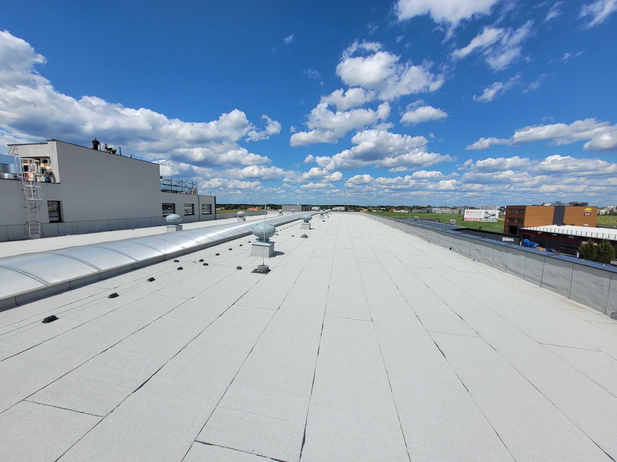 Szeroki, płaski dach przemysłowy pokryty jasną membraną, z widocznymi świetlikami tunelowymi i elementami wentylacyjnymi, na tle błękitnego nieba z chmurami i zabudowań w oddali. Dwóch pracowników...