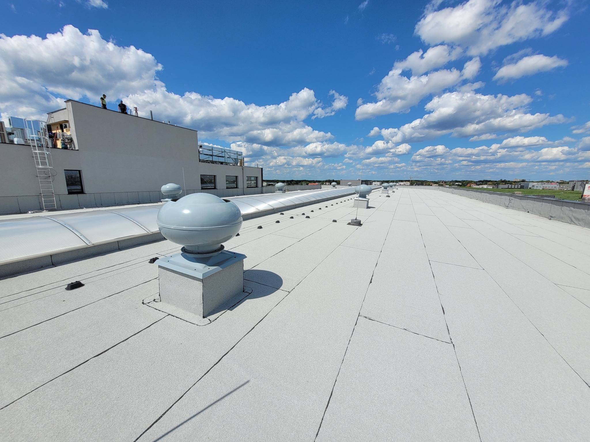 Jasnoszary dach płaski pokryty papą termozgrzewalną z widocznymi kominkami wentylacyjnymi i świetlikami dachowymi, na tle błękitnego nieba z białymi chmurami, w oddali sylwetki dwóch osób...