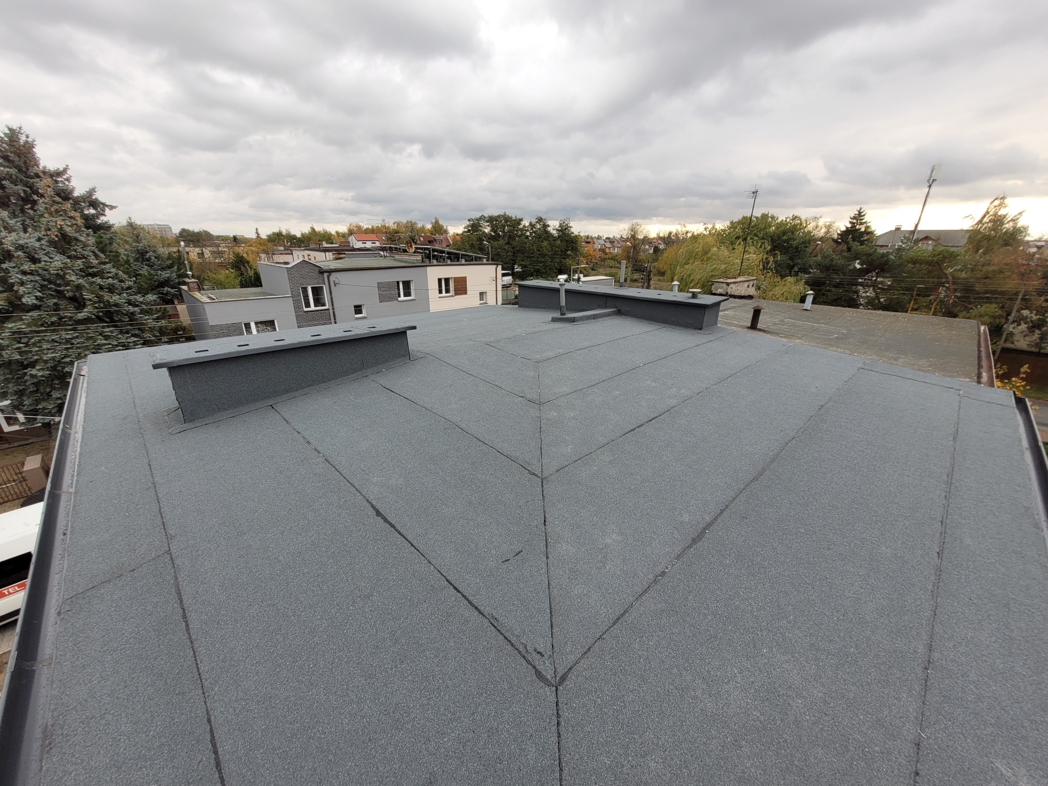 Szary, płaski dach pokryty papą termozgrzewalną z kominami wentylacyjnymi na tle zabudowy miejskiej i pochmurnego nieba. Widok z góry.