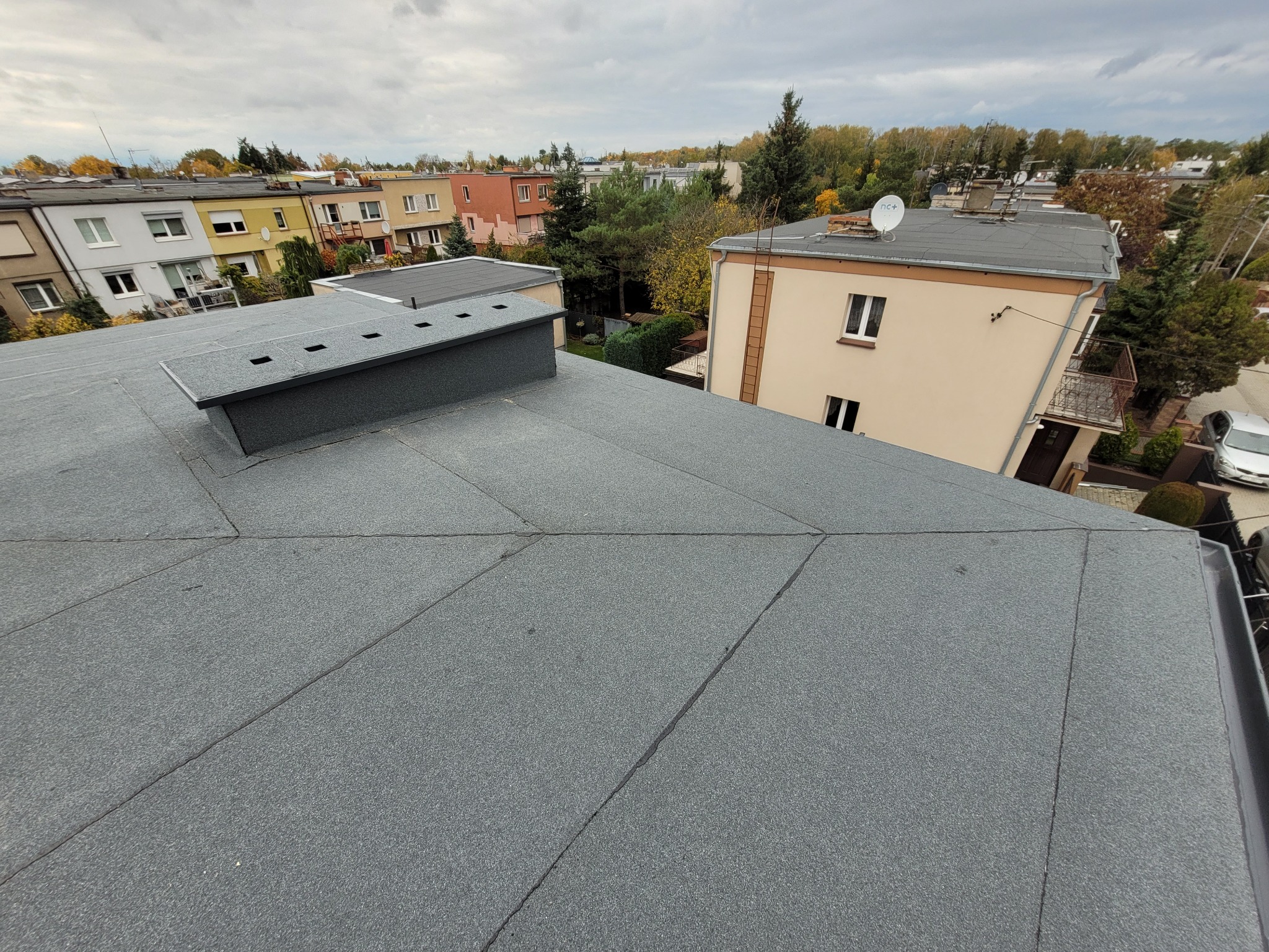 Szary, płaski dach pokryty papą termozgrzewalną z kominem wentylacyjnym. W tle zabudowa jednorodzinna i pochmurne niebo.