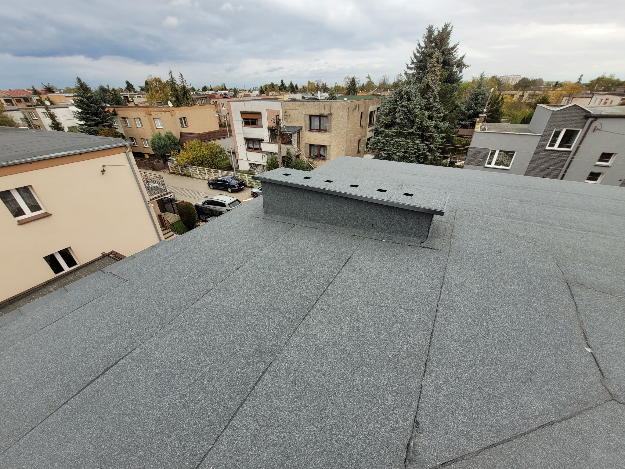 Szary, płaski dach pokryty papą termozgrzewalną z kominem wentylacyjnym na tle zabudowy miejskiej i pochmurnego nieba. Widok z góry, ukośny kadr.