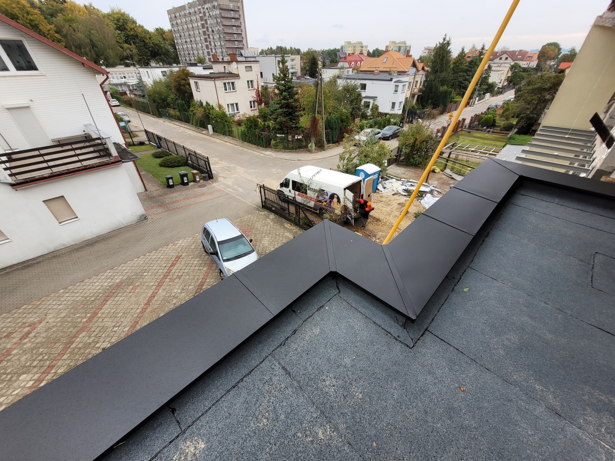 Czarny, płaski dach z obróbkami blacharskimi, widok z góry na tle zabudowy miejskiej i ekipa dekarska przy samochodzie dostawczym z logo firmy.