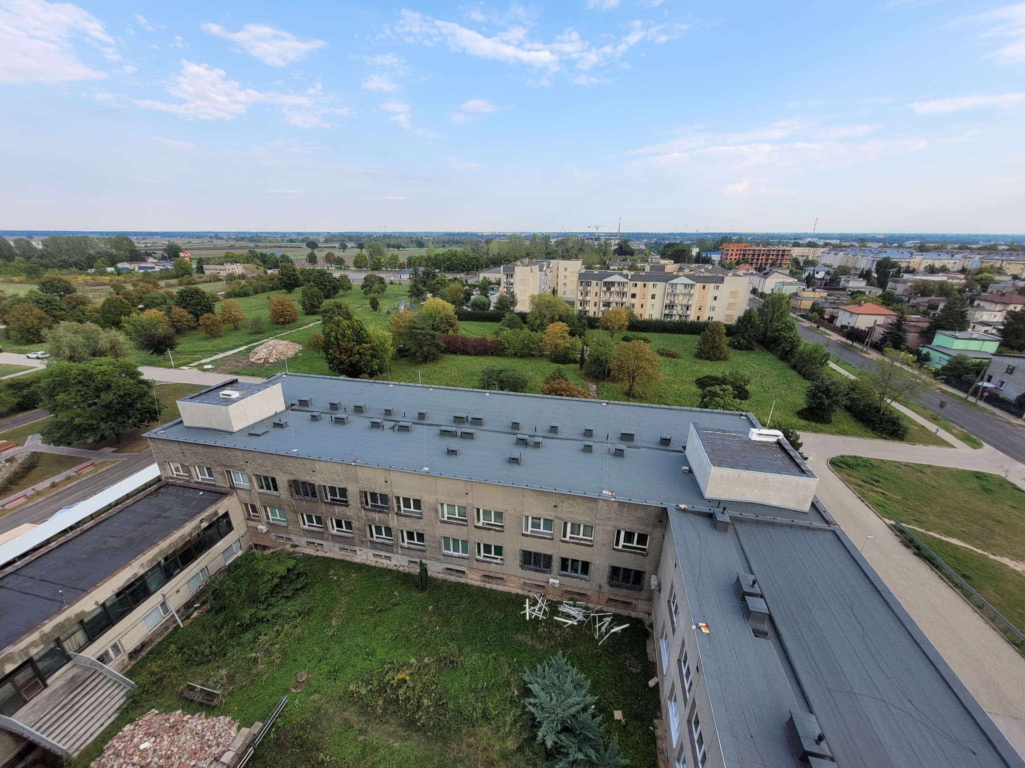 Widok z góry na płaski dach budynku z szarej papy, z licznymi kominami wentylacyjnymi. W tle zabudowa miejska i zieleń. Ujęcie z drona.