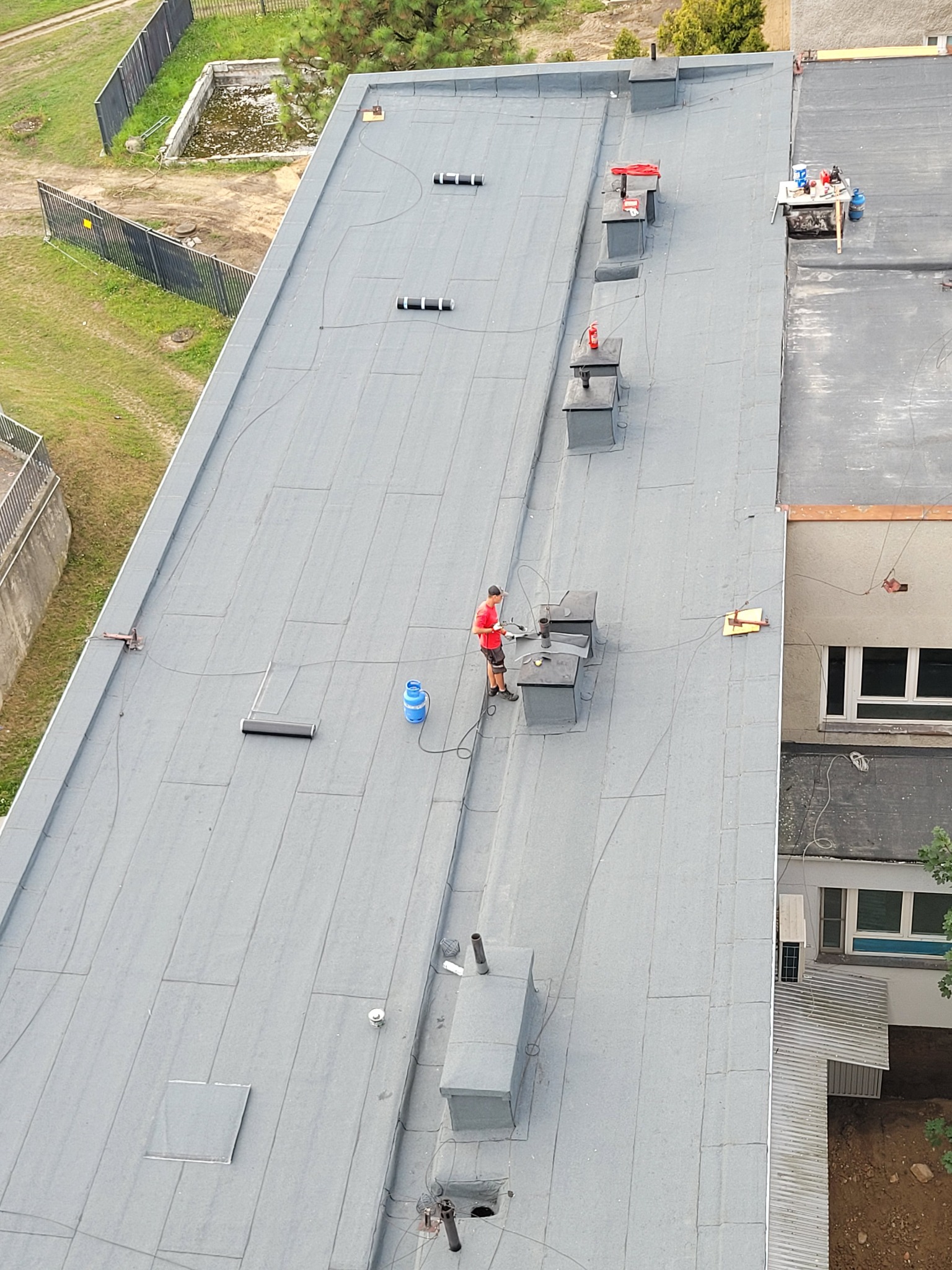 Praca na płaskim dachu budynku: dekarz w czerwonej koszulce przy kominie, obok butla gazowa i narzędzia. Widok z góry, szary dach, elementy wentylacyjne.
