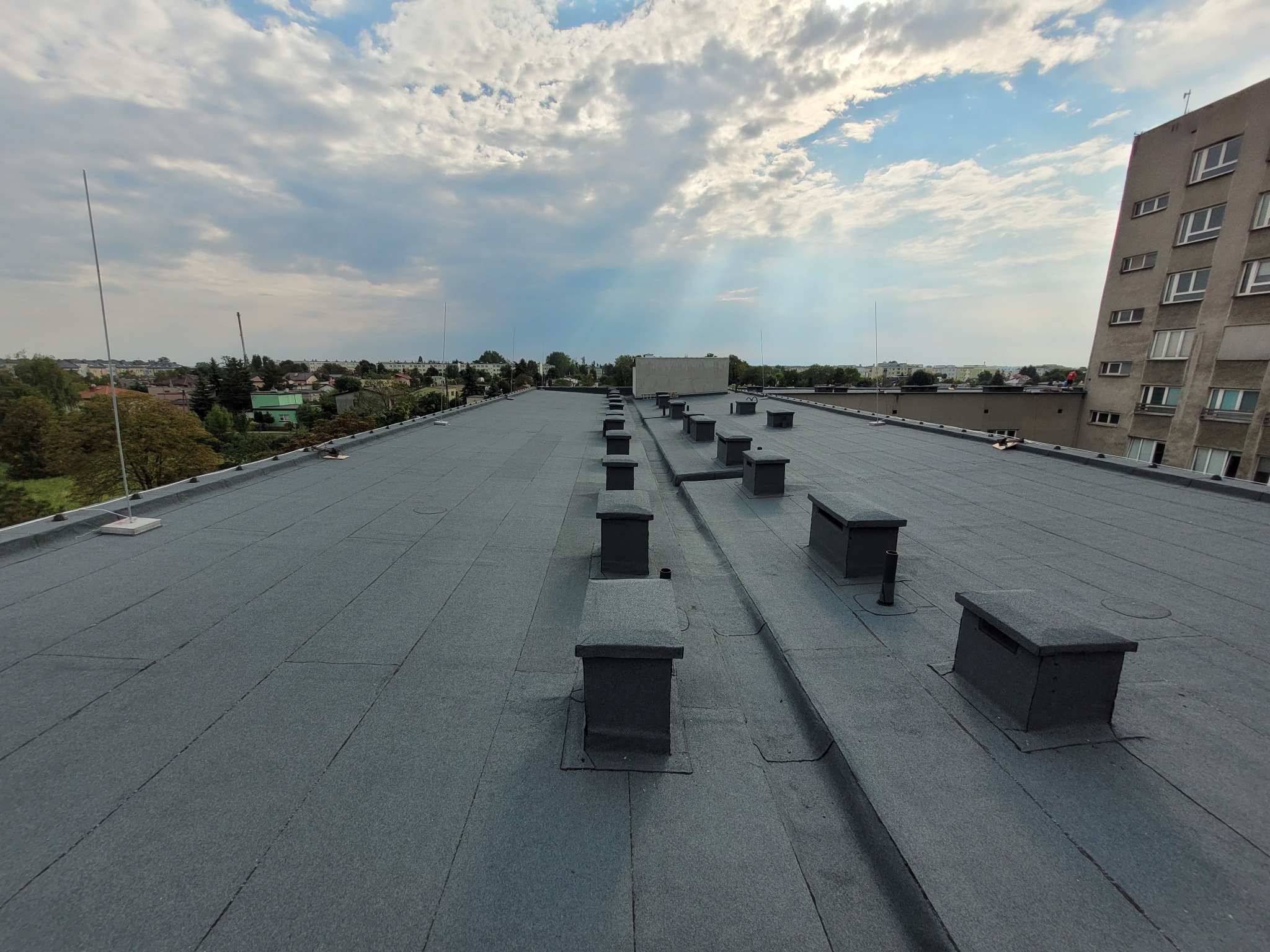 Szary, płaski dach budynku z kominami wentylacyjnymi na tle błękitnego nieba z chmurami. Widok z góry, w tle zabudowa miejska i drzewa.