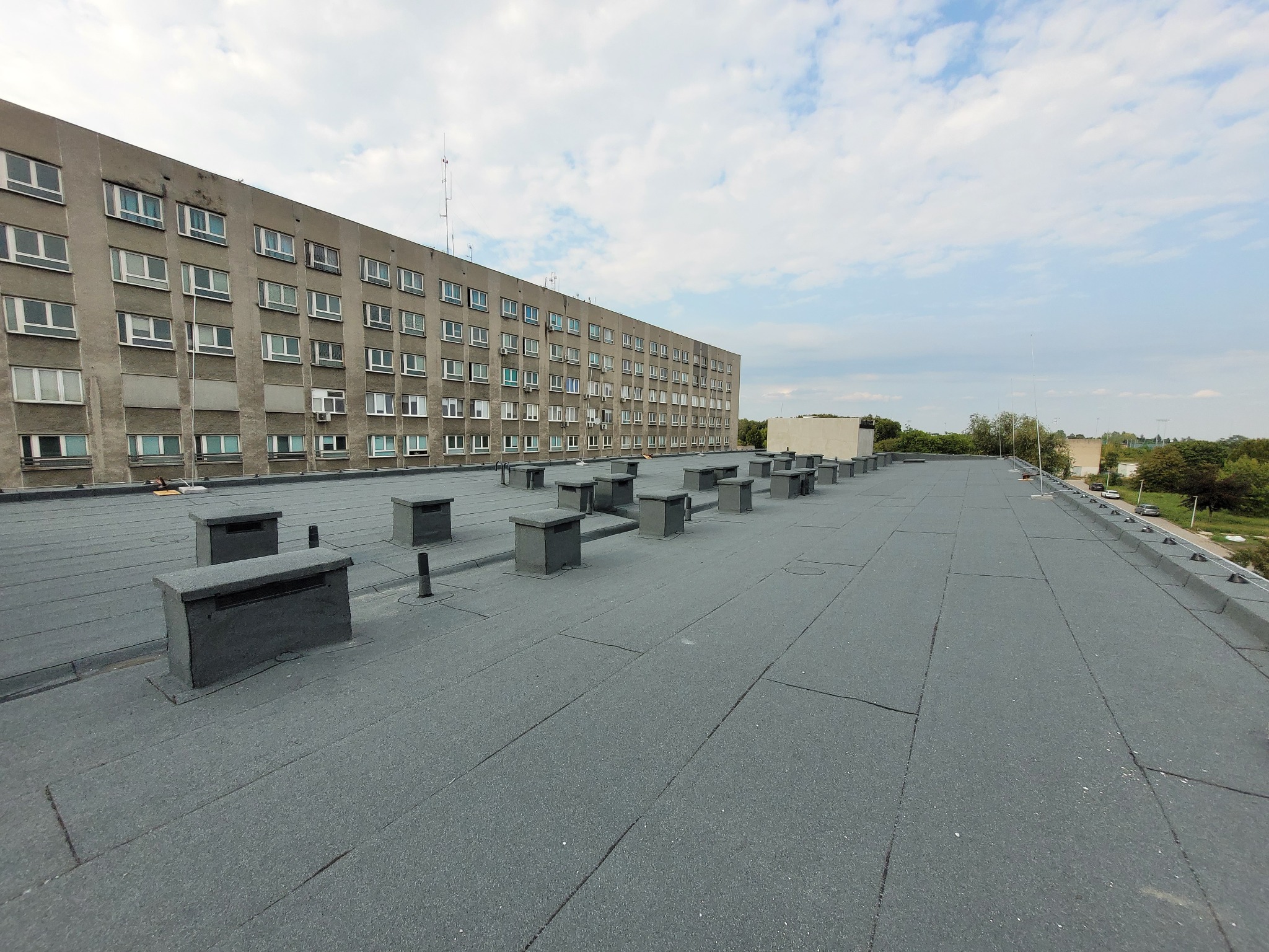 Szary, płaski dach budynku z licznymi kominami wentylacyjnymi. Widok na budynek z wieloma oknami i błękitne niebo z chmurami w tle. Dach pokryty papą.