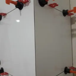 Układanie białych płytek z użyciem systemu poziomowania, widoczne pomarańczowe kliny i czarne podstawy.