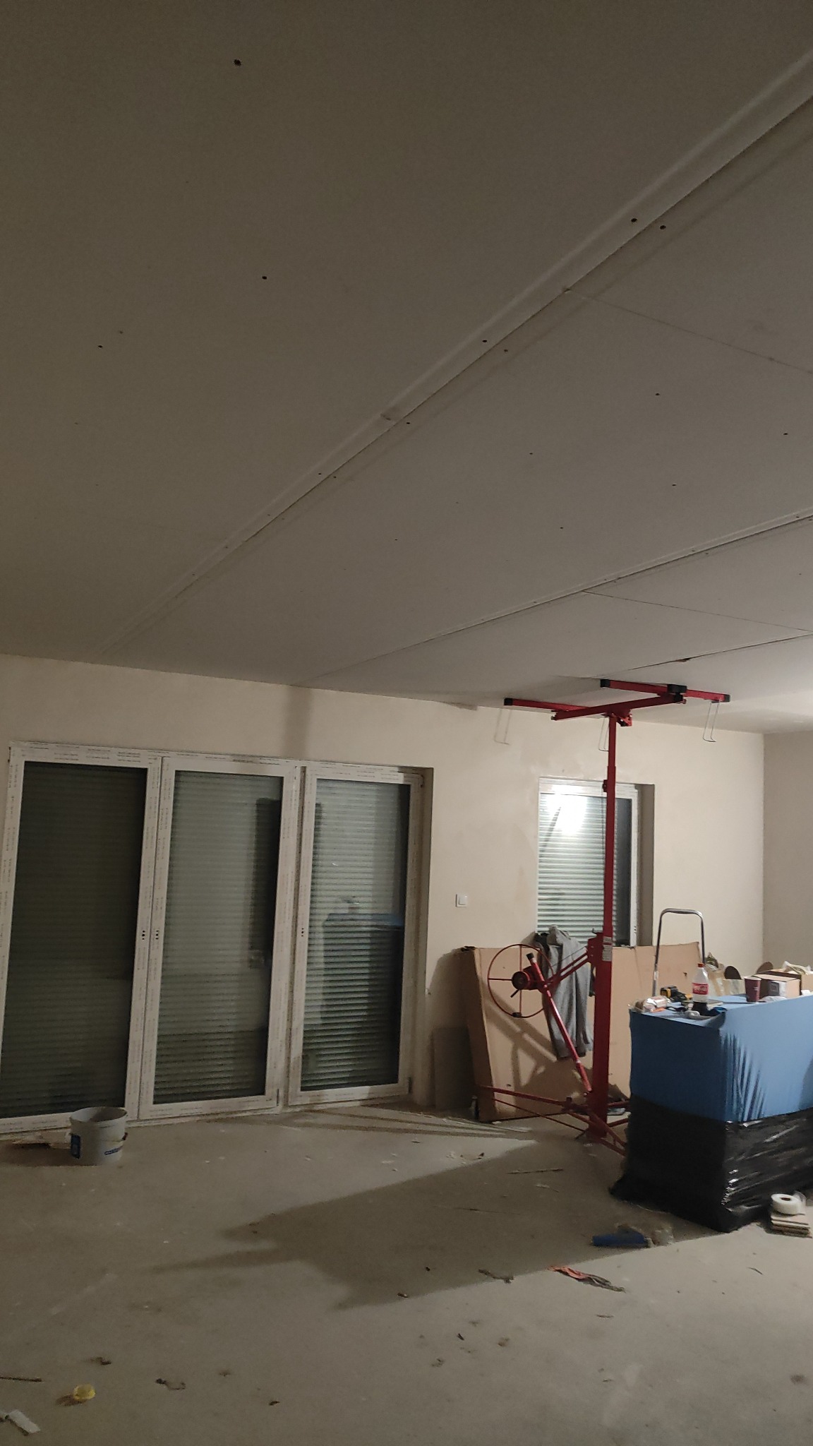 Wnętrze w trakcie montażu płyt gipsowo-kartonowych na suficie, widoczny podnośnik do płyt i częściowo zakryte okno balkonowe.